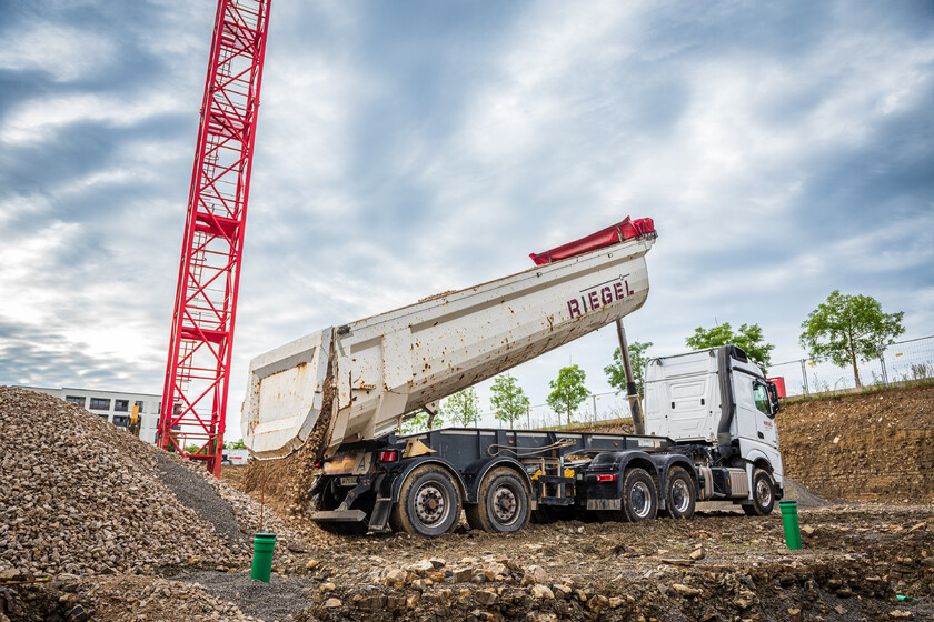 Riegel-Lkw kippt Erdmaterial auf Baustelle neben rotem Baukran bei wechselhaftem Himmel.